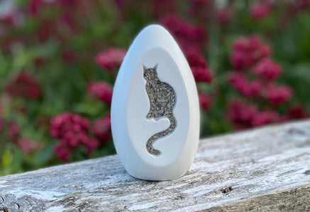 Decorative stone with a cat design on a wooden surface with blurred flowers in the background